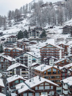 Kışın İsviçre 'nin Zermatt kentinin hava manzarasında kar altında dağ evleri, lüks oteller, modern pansiyonlar, tarla ağaçları, yumuşak bulutlu ışıklar ve düşen karlar görülüyor..