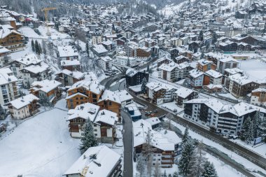 Zermatt, İsviçre 'nin hava manzarası, karlı dağ evi binaları ve otelleri, kıvrımlı nehir ve yolları, Zermatt kilise kulesini, kayak alanlarını, kar küreme makinelerini ve kozalaklı ağaçları gösteriyor..