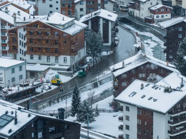 Zermatt, İsviçre 'nin havadan görünüşü, dağ evi çatıları, kıvrımlı bir sokak, donmuş bir nehir, elektrikli araçlar, yayalar, baca buharı ve kışın aktif kar yağışı..