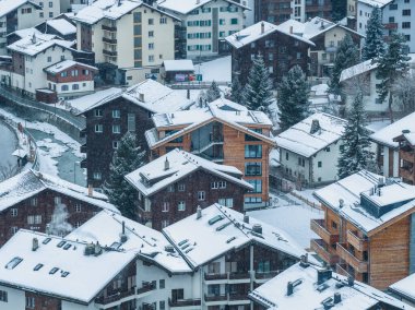 İsviçre, Zermatt 'ın karlı hava manzarasında kısmen donmuş bir nehir kenarındaki dağ evleri ve pansiyonlar, çatılarda ve çam ağaçlarında taze kar, yumuşak bulutlu ışık ve görünür kar yağışı görülüyor..