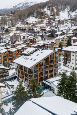 İsviçre 'nin Zermatt kentinin hava manzarasına göre bir yamaçta dağ evi kavgası, derin balkonları, kapalı ve açık havuzu olan bir kereste ve cam otel ve kış ışığı bulutu var..