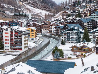 İsviçre 'nin Zermatt kentinin hava görüntüsünde dağ evi tarzı pansiyonlar ve oteller, kompakt sokaklar ve bulutlu aydınlık altında sarp çatılar arasında kıvrılan kanalize bir nehir görülüyor..