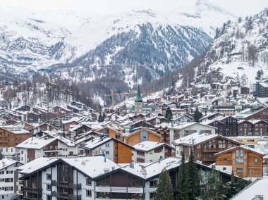 Zermatt, İsviçre 'nin hava manzarası, karlı dağ evleri, Zermatt kilisesinin yeşil kulesi, oteller ve pansiyonlar, çam yamaçları ve gökyüzünü düzleştiren bulutlu ışık gösteriyor..