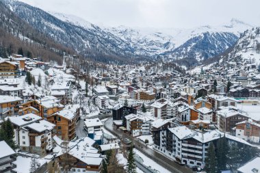 Kışın İsviçre 'nin Zermatt kentinin havadan görünüşü, karla kaplı dağ evleri, lüks oteller, dolambaçlı sokaklar, Zermatt kilise kulesi, kıvrımlı bir yol ve nehir ve alp zirveleri gösteriyor.