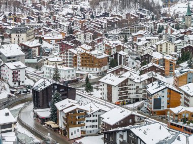 Zermatt, İsviçre 'nin taze karlı havadan görünüşü dağ evleri, modern oteller, dolambaçlı sokaklar, yeşil spiral kilise, turuncu tren vagonları ve bulutlu bir nehirde sergileniyor..