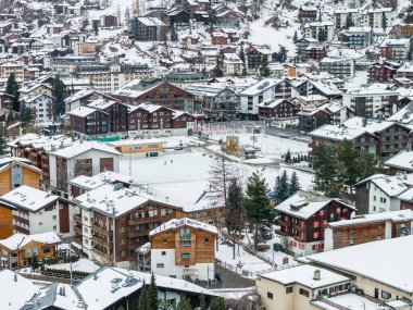 Zermatt, İsviçre 'nin havadan görünüşü, karlı dağ evi kümeleri, açık hava buz pateni pisti, spor alanları, çam ağaçlı sokaklar ve yumuşak bulutlu ışık altında dağlık alanlar..