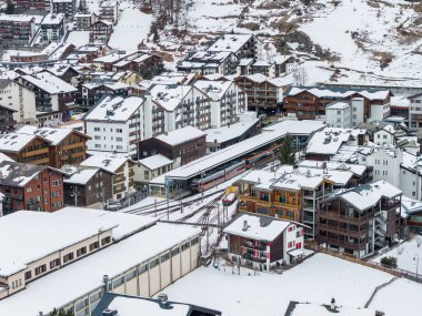 İsviçre 'nin Zermatt kentinin hava manzarası tren istasyonunun etrafındaki kireç ve pansiyonları gösteriyor. Dar ölçü yolları üzerindeki kırmızı ve beyaz vagonlar ve kışın karlı çatılar..