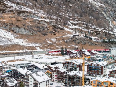 İsviçre 'nin Zermatt kentinin kış manzarası dağ evlerinin çatılarını gösteriyor. Orta kısımda kırmızı bir Matterhorn Gotthard Bahn treni, sarı bir vinç ve sönük ışık yamaçlarında yamalı alak alp yamaçları..