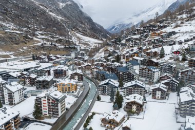 İsviçre 'nin Zermatt kentinin havadan görünüşü, karlı yamaçlardaki Chalet ve oteller, merkezinde dar bir nehir, bulutlardaki dik Alpler ve çatılar arasında bir kilise kulesi..