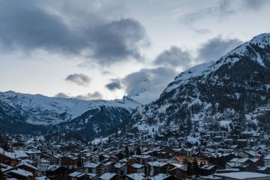 Kış alacakaranlığı Zermatt, İsviçre 'yi gösteriyor. Parlak köy ışıkları, dağ evleri, bulutlardaki Matterhorn, engebeli yamaçlar ve Zermatt kilise kulesi görünür..