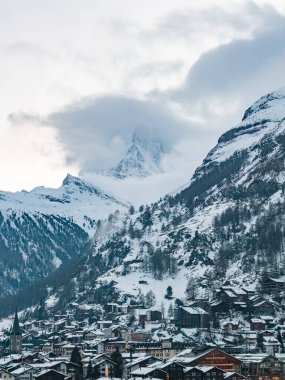 Karlı Zermatt, İsviçre Matterhorn 'un altında bulutlarla örtülü, dağ evleri ve oteller gösteriyor. Zermatt Kilisesi 'nin yeşil kulesi, yumuşak kış günışığında kasabayı demirler..