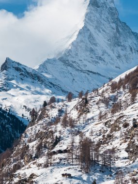 The Matterhorn rises over Zermatt, Switzerland, as mist curls at the summit. Larch dotted slopes descend to rustic wooden chalets under crisp midday alpine skies.