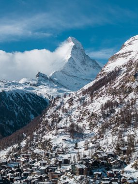 Zermatt, Switzerland sits below the Matterhorn under a crisp sky. Chalet buildings cluster by snow slopes with larch trees in midday light and a wisp of cloud.