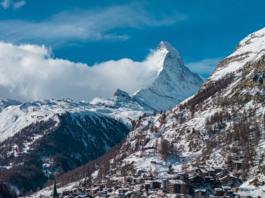 Panoramic view of Zermatt, the Matterhorn, and church spire, with chalets, larch dotted slopes, and sunlit ridgelines under a deep blue sky on a cold clear day.