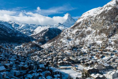 Kışın Zermatt 'ın hava manzarasında dağ evleri, lüks oteller ve kilise kulesi görünüyor. Matterhorn' un ötesine yükselen ışık, ormanlar ve alp yamaçları da var..