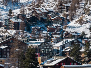 İsviçre 'nin Zermatt şehrinde istiflenmiş dağ evleri ve cam oteller karlı bir yamaca tırmanıyor. Nadir tarlalar yakınlarda duruyor, baca dumanı yükseliyor, ve açık gökyüzü ahşap ve taş dokularını keskinleştiriyor..