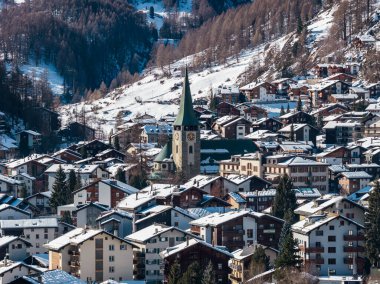 İsviçre 'nin Zermatt kentinin havadan görünüşü, açık kış günışığında kar tozlu dağ başlıklarının ve otellerin ortasında Aziz Mauritius Kilisesi ve saat kulesini gösteriyor..