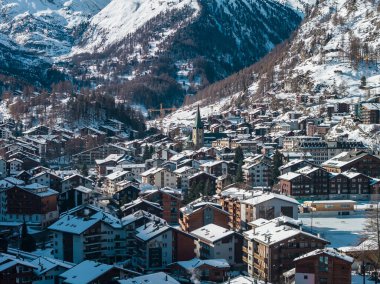 Zermatt, İsviçre 'nin hava öğlen manzarası, kilise kulesi, karlı bardaklar, kayak yamaçları, ağaç tepeleri, düzenli sokaklar ve donmuş bir spor sahası..