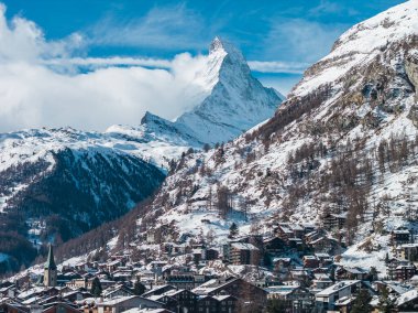 Zermatt, kar çatıları ve yamaçları kaplarken İsviçre Matterhorn 'un altında yer alıyor. Zermatt kilisesinin yeşil kulesi oteller ve kiraz ağaçlarının arasından gün ışığında yükselir..