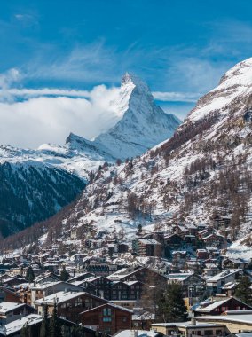 Zermatt, İsviçre Matterhorn 'un altında oturuyor. Karlar çatıları, kilise kuleleri, çam noktalı yamaçlar, gün ışığı parlıyor, tepenin etrafında bulutlar kıvrılıyor..