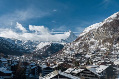 Matterhorn 'un altındaki Zermatt, İsviçre' de kar temizleme bezleri. Merkezde bir kilise kulesi yükselir, oteller yamaçları katlar, ve gün ortası ışığı açık bir kış gökyüzünü tanımlar.