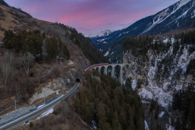 Hava görüntüsü, Filisur yakınlarındaki Landwasser Viaduct 'tan geçen kırmızı Buzul Ekspresi' ni gösteriyor, İsviçre Alpleri, alacakaranlıkta, çam ormanları, kireçtaşı duvarlar ve yamaçlarda kar..