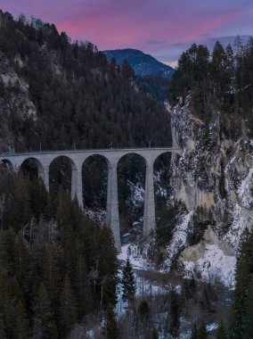 Yüksek bir manzara Graubunden, İsviçre 'deki Landwasser Viaduct' u gösteriyor. Buzul Ekspresi mor bir gökyüzünün altındaki uçurum tüneline girerken altı kireçtaşından kemer var..