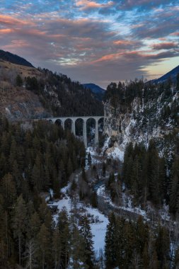 Hava görüntüsü Filisur, Graubunden, İsviçre yakınlarındaki karlı bir Alp geçidine uzanan Landwasser Viaduct 'ı gösteriyor. Nehir, kozalaklı ağaçlar, pembe bulutlar ve Buzul Ekspres yolu..