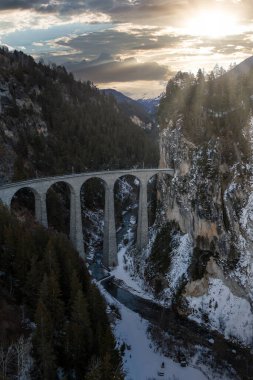 Hava görüntüsü Filisur, İsviçre Alpleri yakınlarındaki Landwasser Viaduct 'u gösteriyor. Landwasser Nehri 'nin üzerindeki ormanlık bir uçurumdan altı kavis. Gün batımı ışınları kışın kayaları ve buzları aydınlatıyor..
