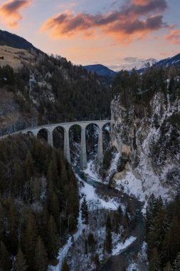 Hava kararma sahnesi, Graubunden, Filisur yakınlarındaki Landwasser Viaduct 'u gösteriyor. Taş kemerler, kar çamları ve sıcak pembe bulutlarla dolambaçlı buzlu bir nehrin üzerinde bir vadi boyunca uzanır..
