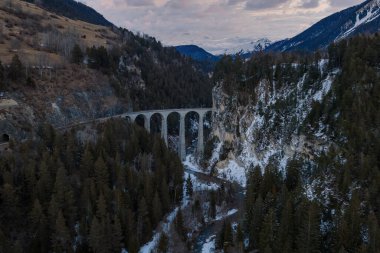 Hava görüntüsü, Filisur, Graubunden yakınlarındaki derin bir geçitten geçen Landwasser Viaduct Viaduct 'un altı taş kemer, çam ormanı, nehir ve bulutların altında karlı zirveler olduğunu gösteriyor..
