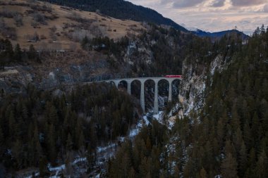 Gökyüzü görüntüsü Graubunden, İsviçre 'deki Landwasser Viaduct' u gösteriyor. Parlak kırmızı Buzul Ekspresi kışın sonlarında bir orman geçidinde altı taş kemer geçiyor..