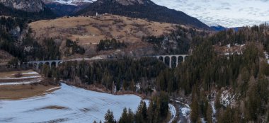 Hava görüntüsü, Graubunden 'da bir orman geçidinde kavis çizen Landwasser Viaduct' ı gösteriyor. Taş kemerler, kar tozlu yamaçlar, köyler, nehir ve yamalı kış tarlaları..