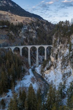 Graubunden, İsviçre 'deki Landwasser Viyadük' ünün hava görüntüsü, altı taş kemer kıvrımlı donmuş bir nehrin, dik orman kayalıklarının ve kar tozlu yamaçların üzerinde bir vadi boyunca uzanıyor..