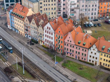 Aerial view of pastel gabled townhouses in Wroclaw, Poland, near the New European Center, as a blue tram approaches, pedestrians walk, and parked cars sit behind