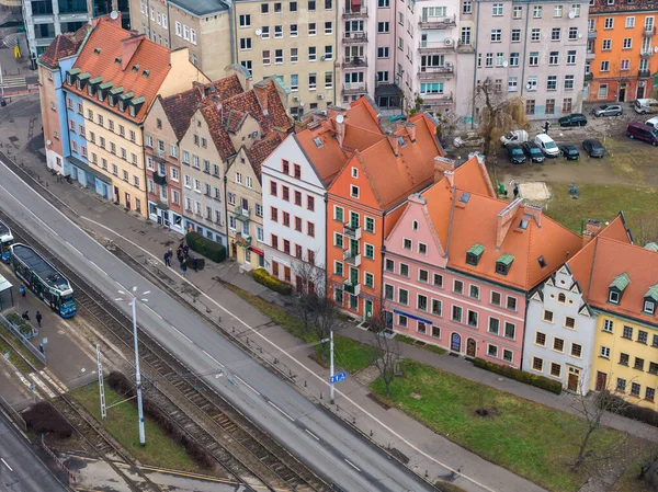 Aerial view of pastel gabled townhouses in Wroclaw, Poland, near the New European Center, as a blue tram approaches, pedestrians walk, and parked cars sit behind
