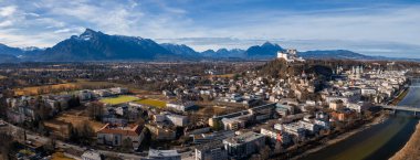 Aerial view of Salzburg, Austria, with Hohensalzburg Fortress above the old town, the Salzach River on the right, baroque spires, bridges, and the snow dusted Alps at midday.