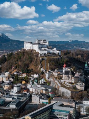 Avusturya, Salzburg 'un hava manzarası Festungsberg' deki Hohensalzburg Kalesi 'ni gösteriyor. Pastel evler, yeşil bakır kubbeler, kırmızı kule ve bulutlu gökyüzü altındaki Alp zirveleri..