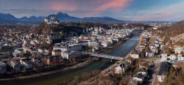 Aerial view of Salzburg, Austria, with the Salzach River, multiple bridges, and Hohensalzburg Fortress. Baroque Salzburg Cathedral rises as Alps sit distant under winter daylight.