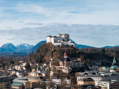 Aerial Salzburg, Austria, shows Hohensalzburg Fortress above the old town, green copper domes, a red spired clock tower, and snow dusted Alps in cool even daylight.