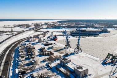 Kışın Riga Tersanesi 'nin hava manzarası Daugava Nehri' ndeki buzlu rıhtımları, mavi kanat vinçlerini, demirlemiş gemileri, kuru rıhtımları, karlı bahçeleri ve parlak gün ortası gökyüzünü gösteriyor..