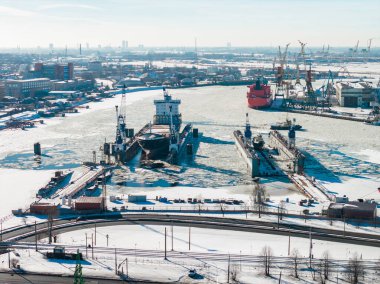 Riga Tersanesi 'nin donmuş Daugava Nehri üzerindeki havadan görünüşü, kuru rıhtımlar, vinçler, kırık buzlar, sağa demirlemiş kırmızı bir kargo gemisi, demiryolu rayları, soğuk hava, Riga Limanı..