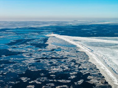 Hava sahnesi Riga 'daki donmuş Daugava Nehri girişini gösteriyor. Kırmızı Mangalsala deniz feneri karlı bir dalgakıran, buz kütleleri, aylaklar ve iki kargo gemisi..