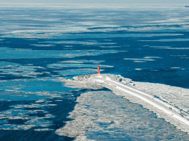 Riga, Letonya 'daki Mangalsala Deniz Feneri' nin havadan görünüşü. İnsanlar buzda yürürken, kırmızı bir kule karla kaplı bir dalgakıranla yükseliyor. Riga Tersanesi yakınlarında mavi gri buz sürükleniyor.