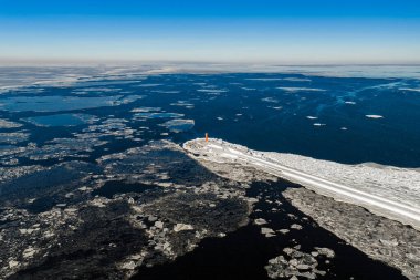Letonya Riga 'daki Daugava Nehri ağzındaki Mangalsala Deniz Feneri' nin havadan görüntüsü. Açık gökyüzünün altında karla kaplı bir dalgakıranı çevreleyen buz salları ve kırık yüzgeçler..