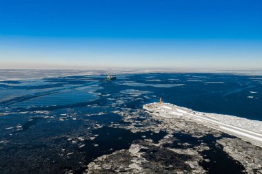 Havadan bakıldığında Mangalsala Deniz Feneri, Daugava Nehri 'nin ağzında buzla kaplı bir dalgakıran, büyük dalgalar ve karanlık kablolar ve Riga Limanı' na doğru giden bir kargo gemisi kanalı görülüyor..