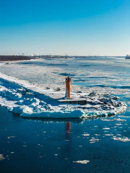 Letonya, Riga 'da Baltık Denizi' yle buluşan donmuş Daugava Nehri 'nin havadan görüntüsü. Mangalsala Deniz feneri kayalık bir iskelenin üzerinde buzlu, iskele vinçleri, sürüklenen deniz buzu ve karanlık su..