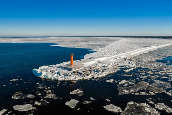 Hava sahnesinde donmuş Mangalsala Deniz feneri Riga, Letonya 'daki Daugava nehrinin ağzında donmuş bir dalgakıran üzerinde yüzen dalgalar ve kış güneşi altında kobalt denizi görülüyor..