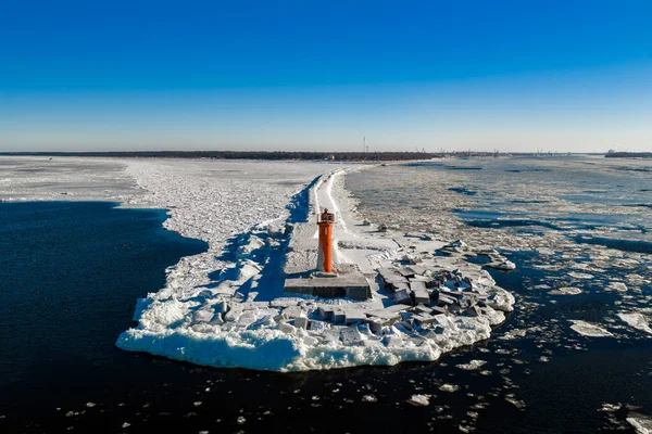 Letonya Riga 'da, buz kabuklu bir dalganın üzerindeki parlak turuncu Mangalsala Deniz Feneri' nin havadan görünüşü. Kırık deniz buzu, karanlık su, tetrapodlar ve tersane turnaları görülebilir..