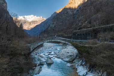 Taş kemer köprüsü, İtalyan Alpleri 'ndeki bir turkuaz akıntısını kapsar. Köprüde bir kişi duruyor. Öğleden sonra kar zirveleri parlar. Bir tünel duvarı kemerli açıkları gösteriyor.